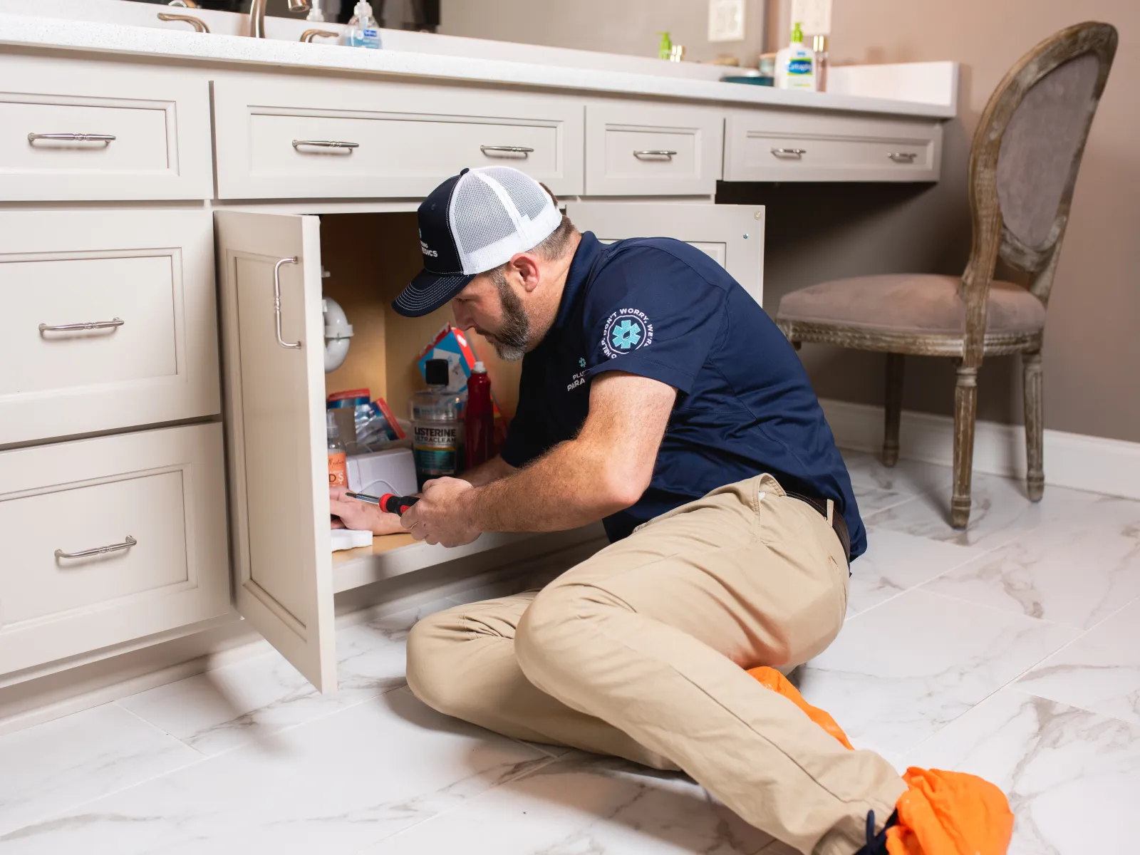 Technician in uniform repairing plumbing under bathroom sink cabinet with tools on white tiled floor.