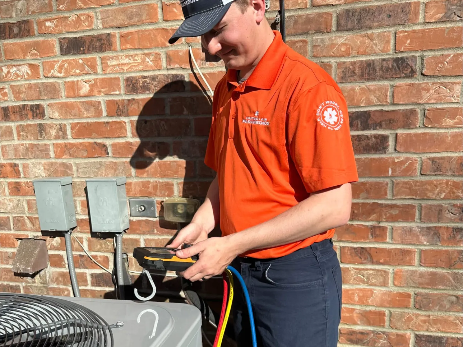 Technician in orange shirt servicing air conditioning unit outdoors with diagnostic tools against brick wall.