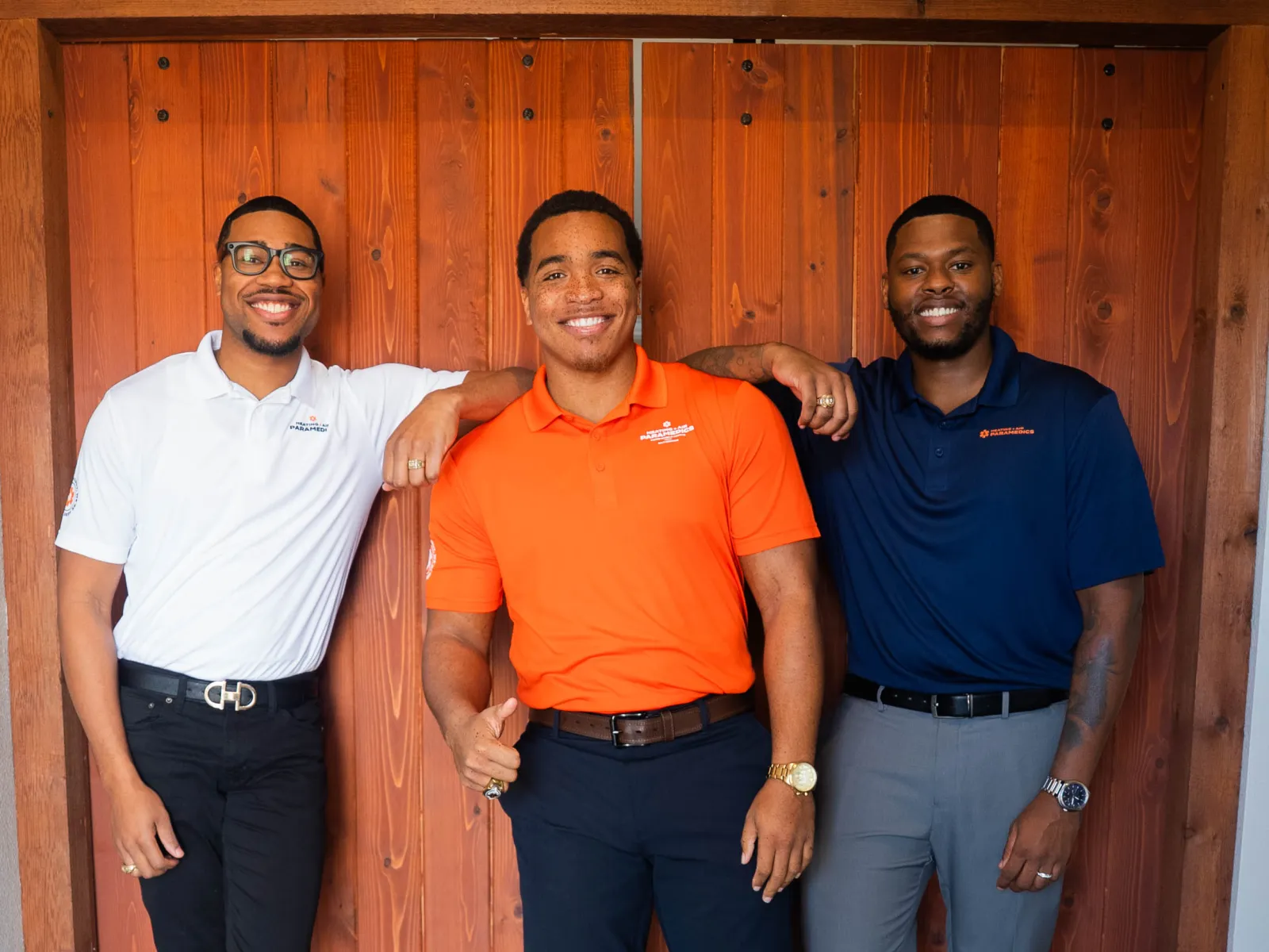 Three smiling men in colorful polos standing in front of a wooden door on dark hardwood floor indoors.