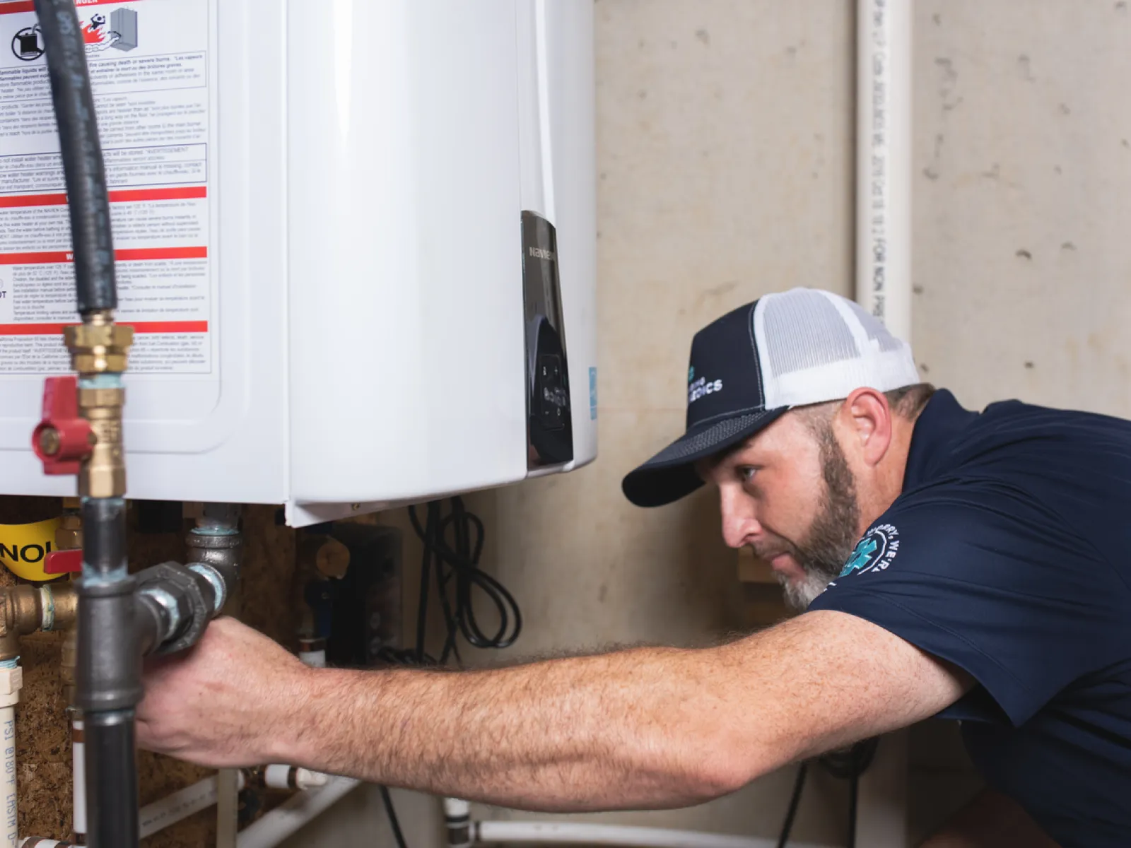 Technician wearing cap inspects and adjusts pipes on a tankless water heater in a basement setting.