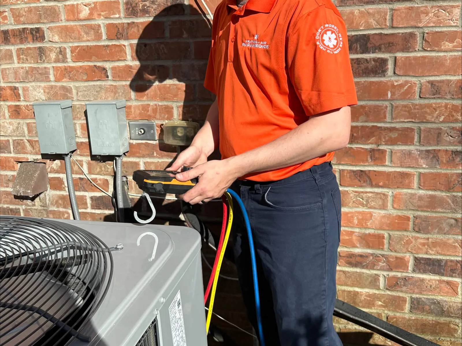 Technician in orange shirt and cap using tool to service outdoor air conditioning unit against brick wall.