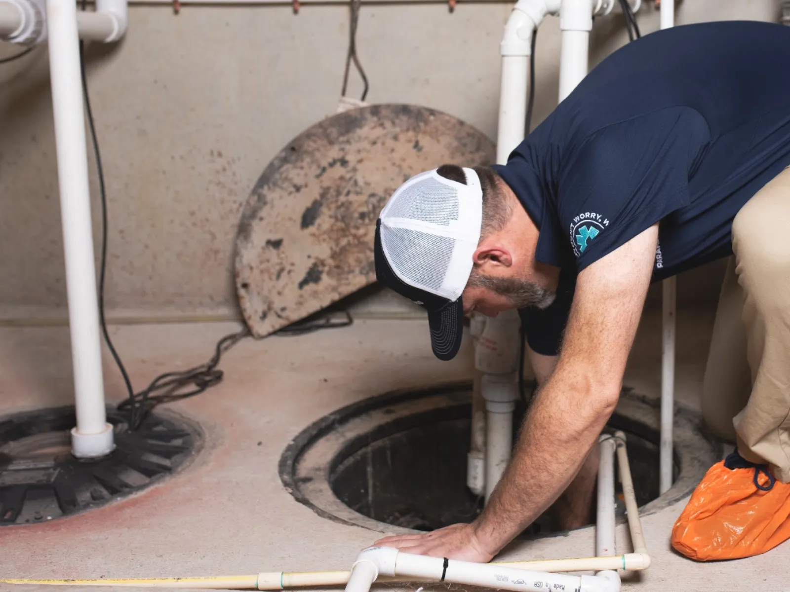 Technician inspecting and working inside a sump pump basin with white plumbing pipes in a basement.