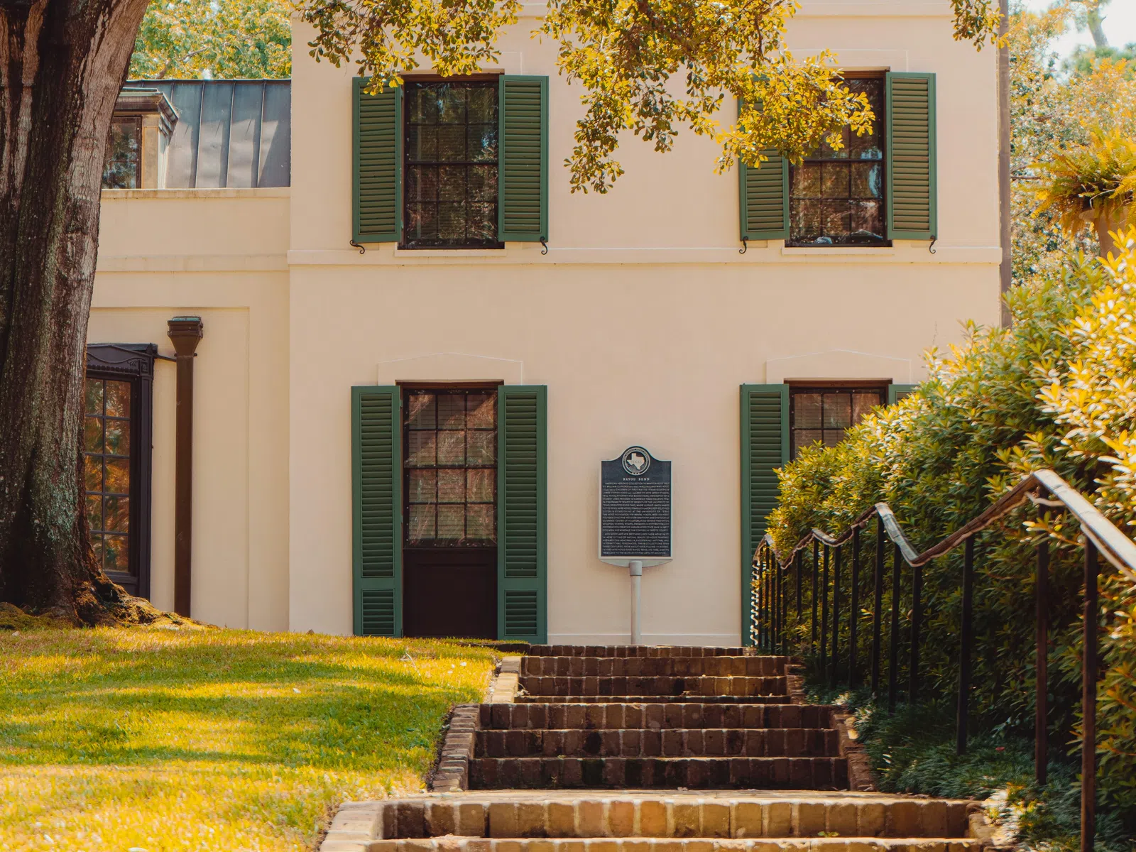 Brick steps leading to a cream-colored historic building with green shutters and a large tree overhead.