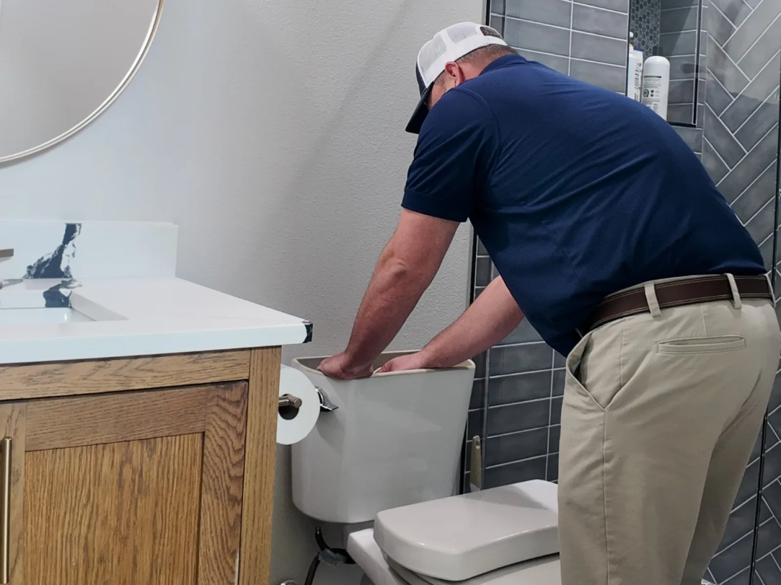 Man in blue shirt fixing or inspecting a toilet tank in a modern bathroom with patterned tile floor and glass shower