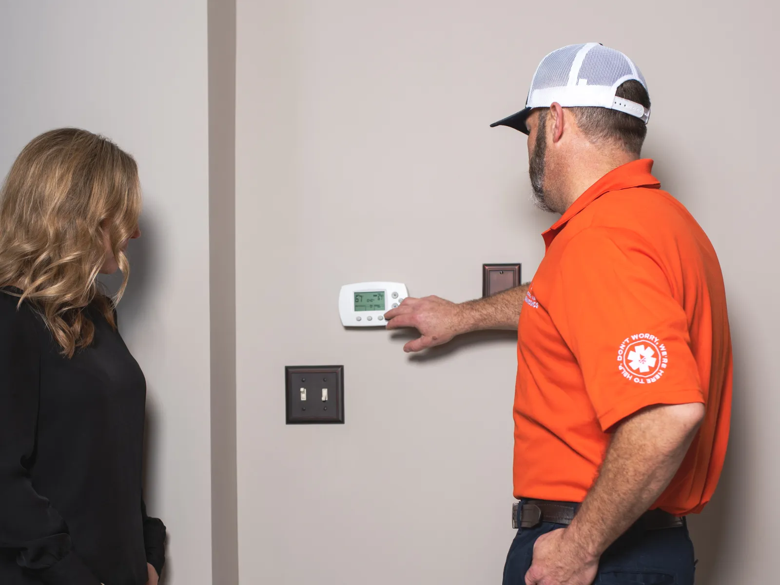 Technician in orange shirt adjusting thermostat on wall while woman watches in a home interior.