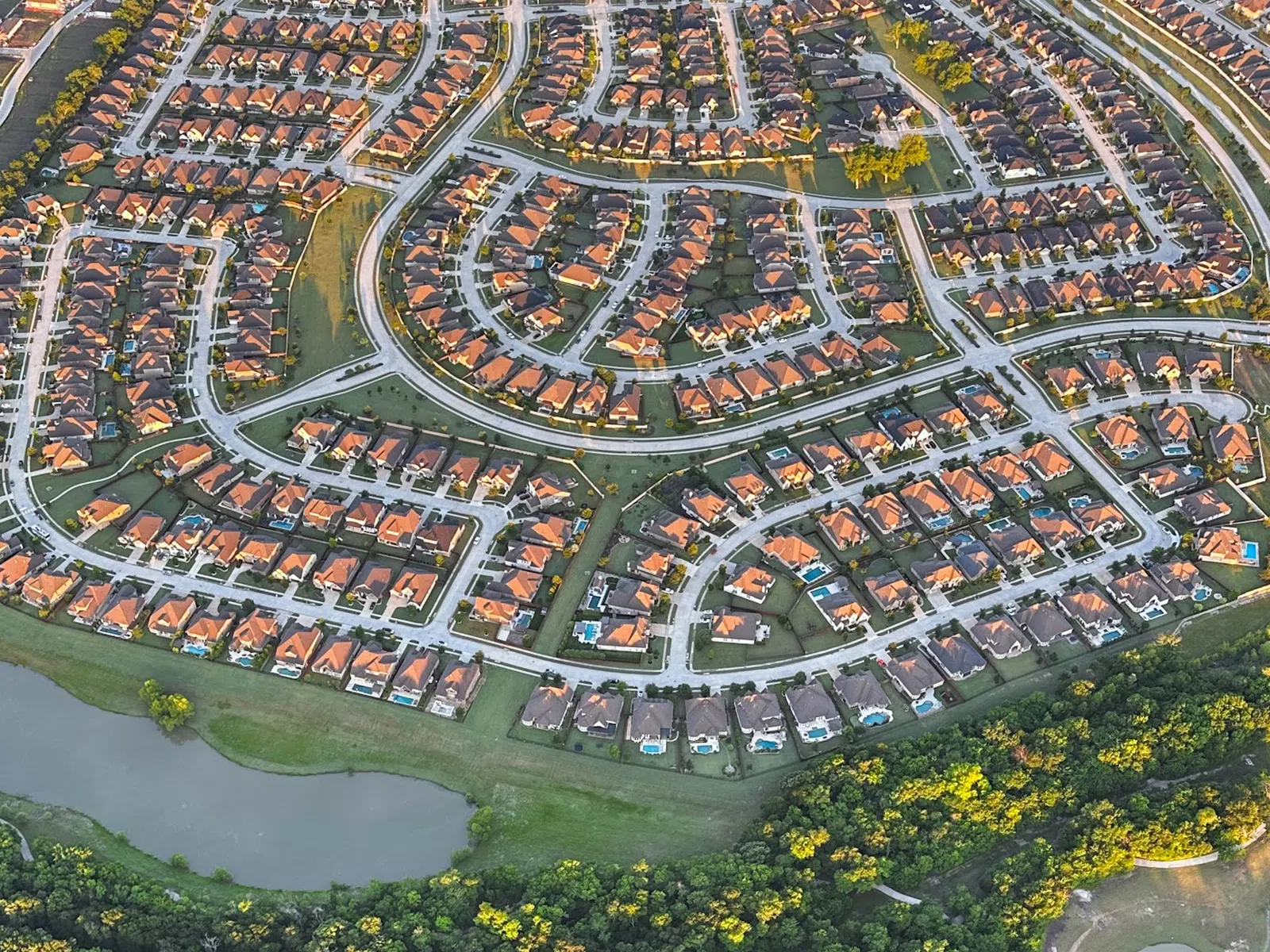 Aerial view of a suburban neighborhood with houses and swimming pools adjacent to a dense forest and a golf course.