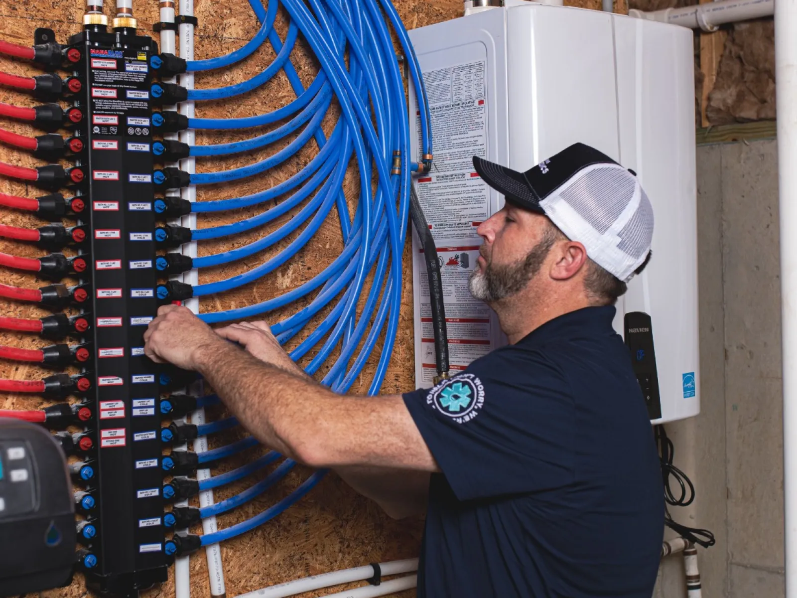 Technician adjusting plumbing manifold with blue and red tubes in a utility room near a white tankless water heater.