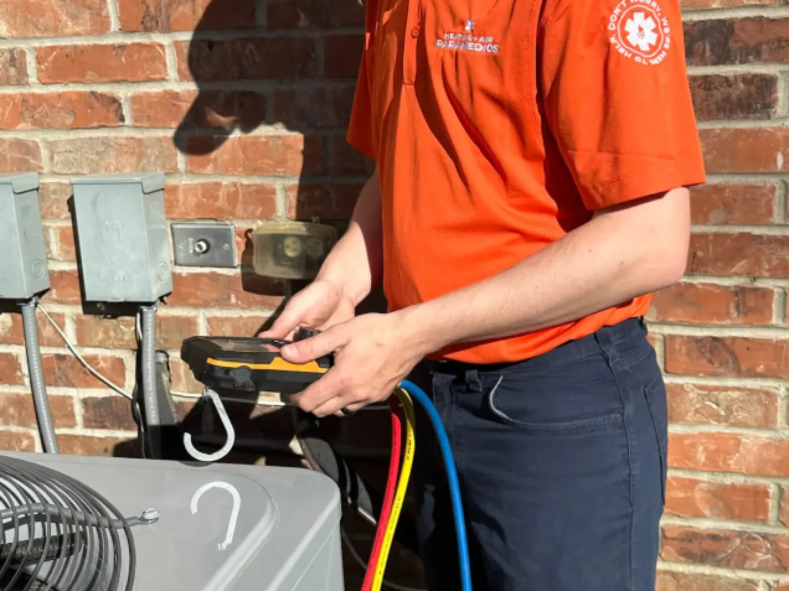 Technician in orange shirt using device to check HVAC unit outdoors against brick wall.