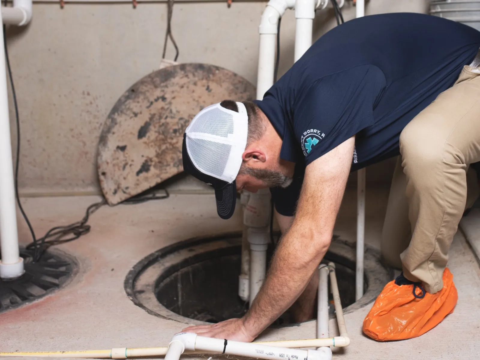 Technician inspecting and working inside a sump pump pit in a basement with plumbing pipes around.