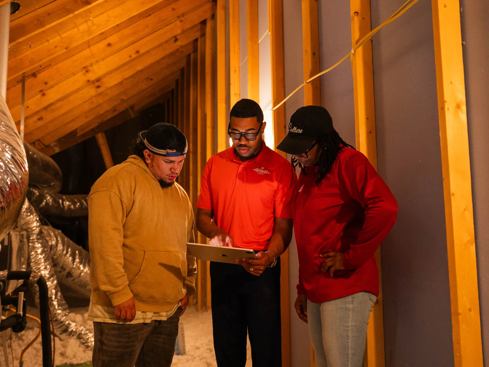 Three workers examine a tablet in an attic under construction with exposed wood framing and ducts.