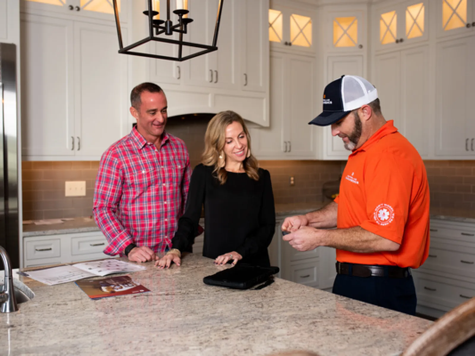 Technician in orange shirt explaining details to couple in modern kitchen with white cabinets and granite countertop.
