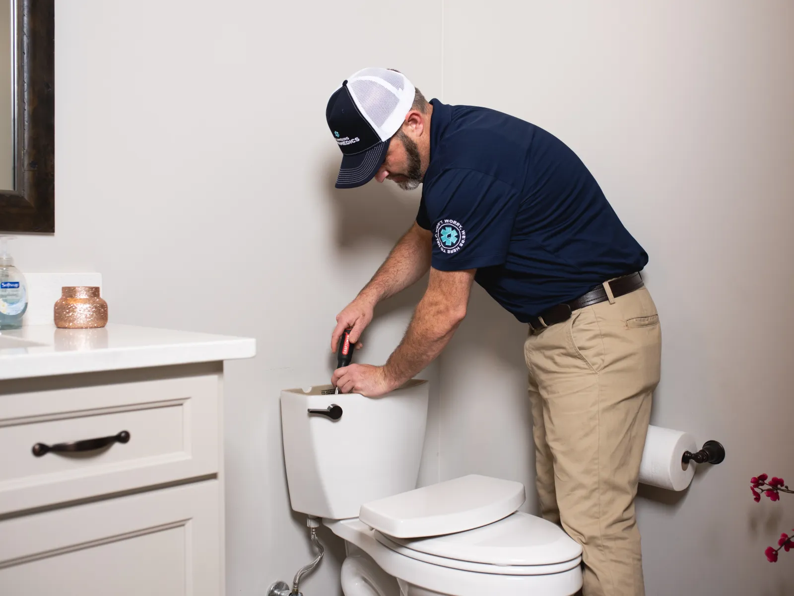 Plumber in uniform repairing a toilet tank in a bathroom with beige walls and white cabinetry.