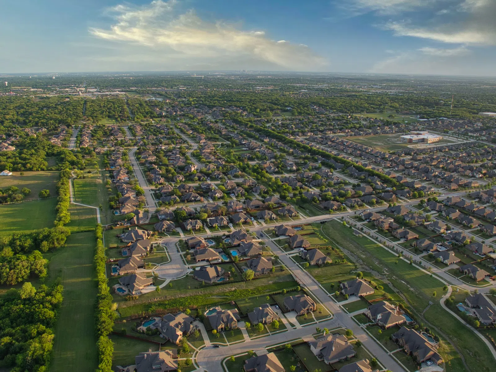 Aerial view of a sprawling suburban neighborhood with houses, roads, and green spaces under a partly cloudy sky.