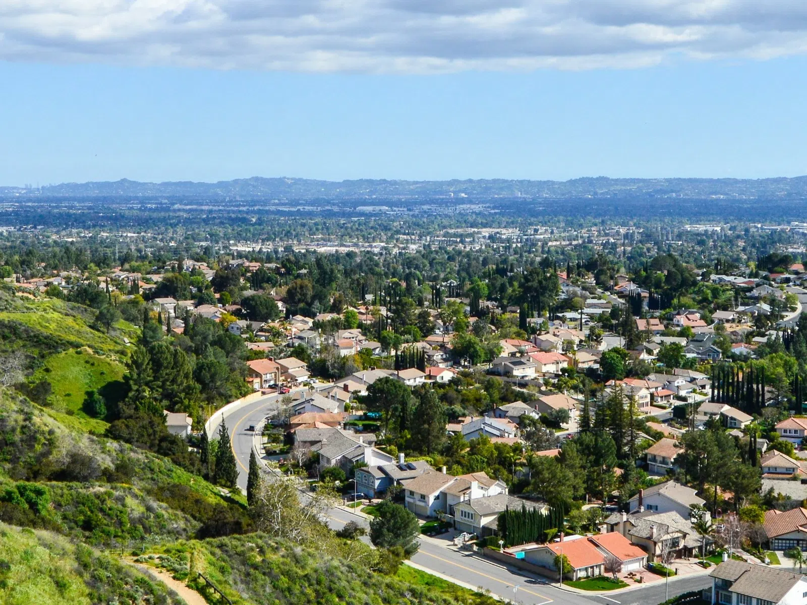 Panoramic view of a suburban neighborhood with houses, trees, winding roads, and distant hills under a partly cloudy sky