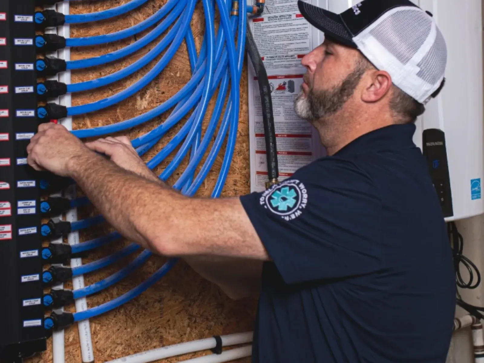 Technician adjusting blue PEX tubing in a plumbing manifold system mounted on a wooden board indoors.