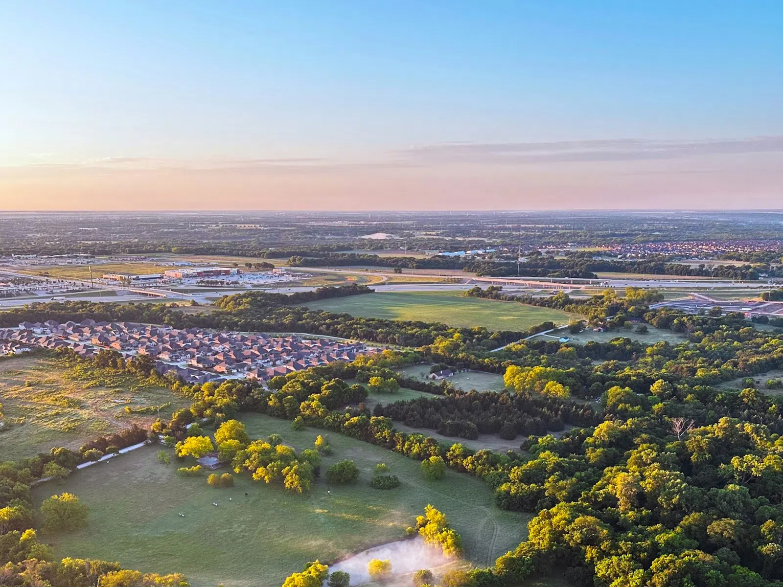Aerial view of lush green forest with scattered clearings and a distant suburban neighborhood under clear blue sky.