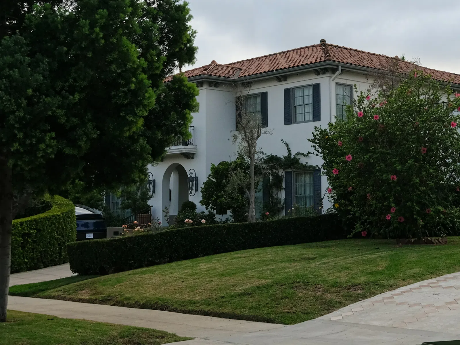 White two-story house with red tile roof surrounded by trees and bushes on a cloudy day.