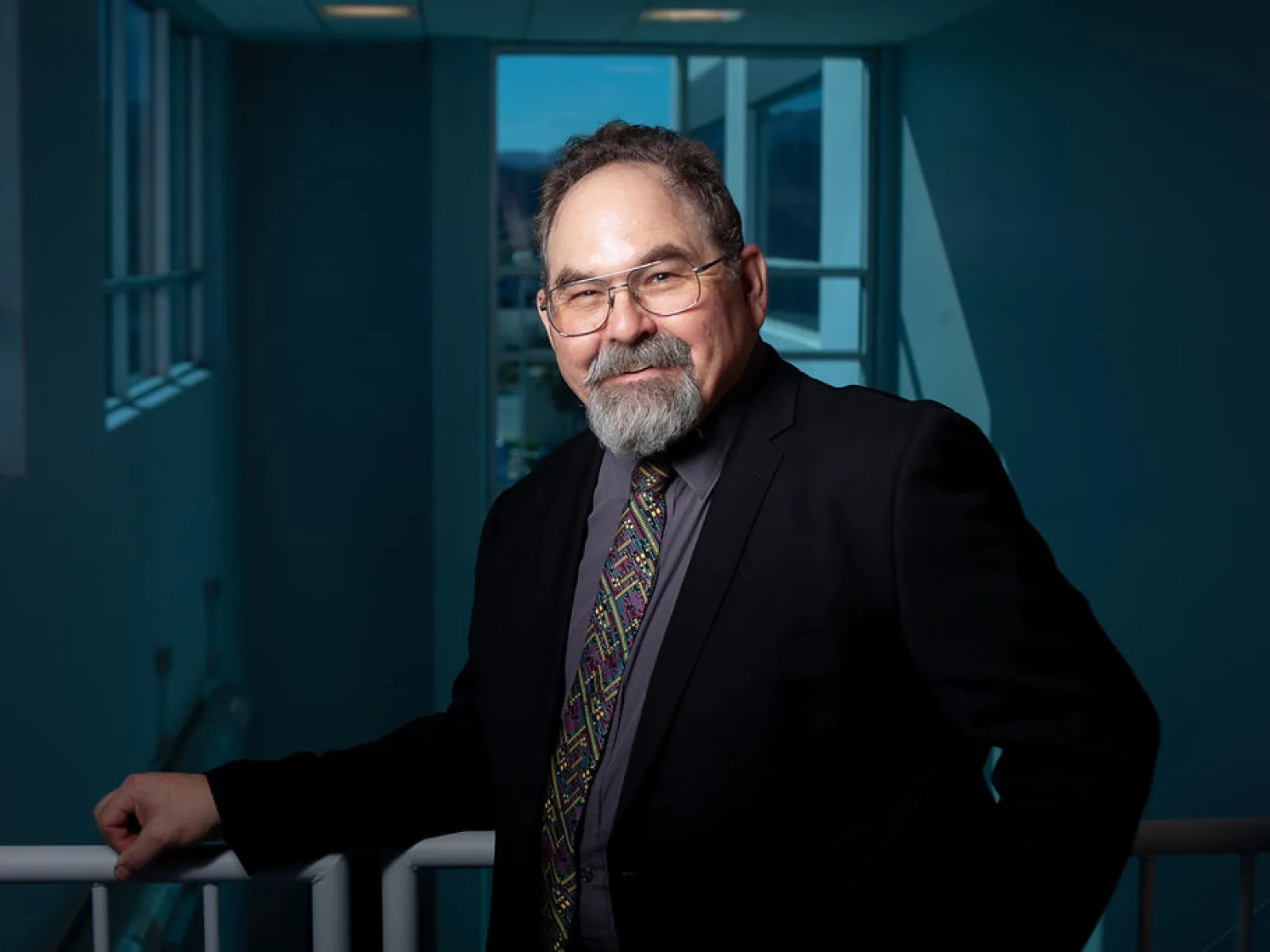 Mature man in glasses and patterned tie smiling confidently against a modern blue-toned indoor background.