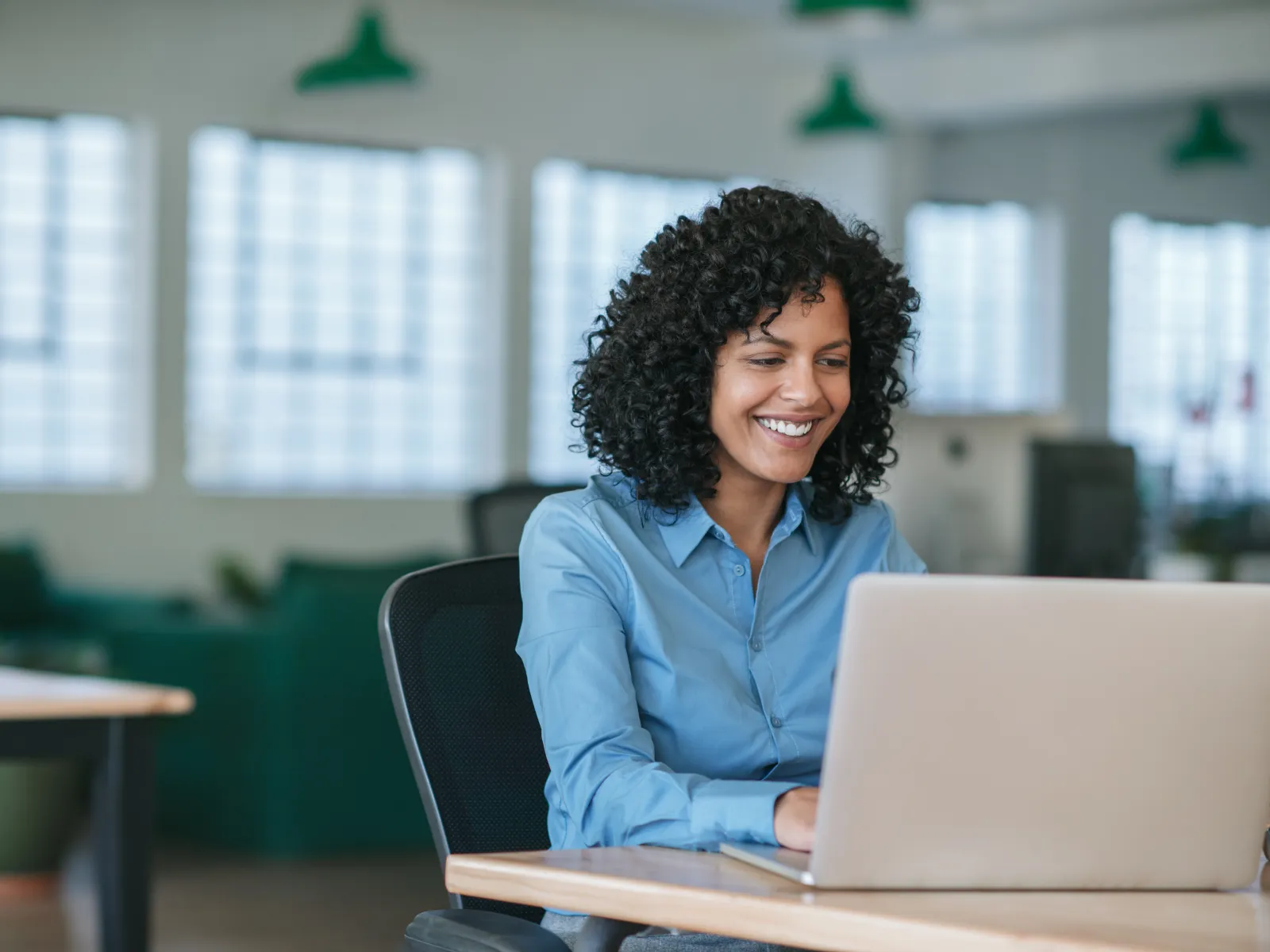 Young woman with curly hair works happily on a laptop in a bright modern office space with large windows.