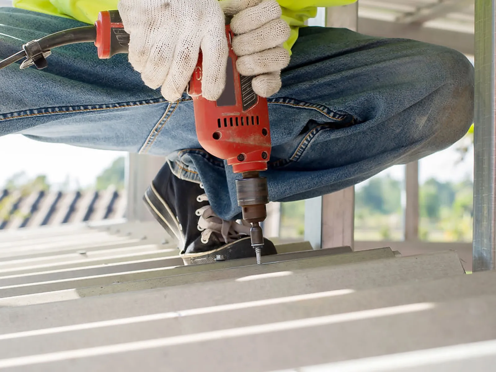 Construction worker in gloves using electric drill to fasten screws on metal roofing under sunlight.