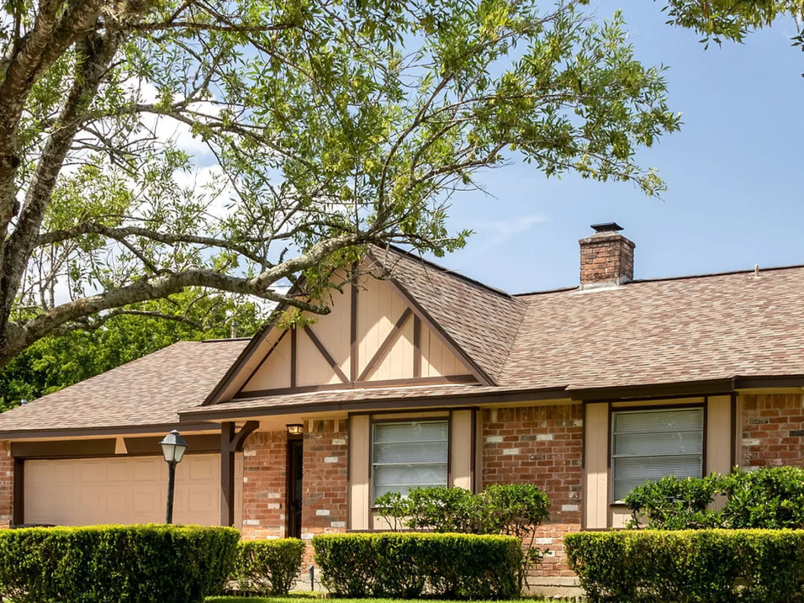 Brick suburban house with large green lawn, trimmed hedges, and leafy tree branch on a sunny day.