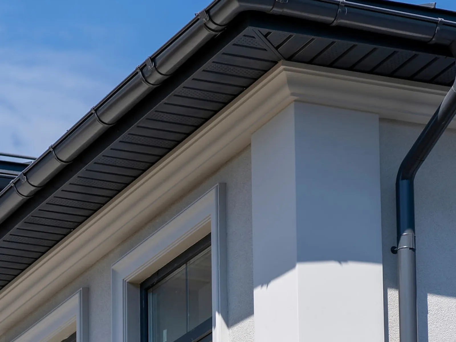Close-up of modern white house corner with windows and black rain gutter under blue sky.