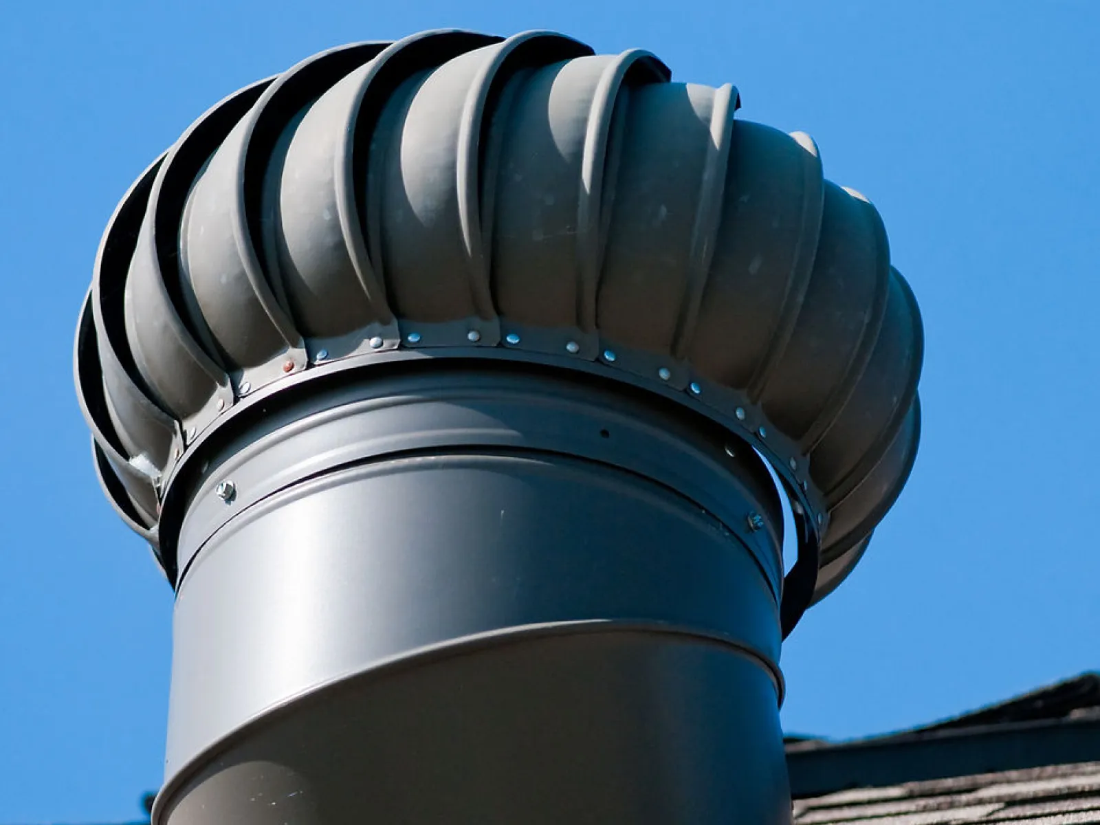 Close-up of a metal roof turbine vent installed on shingled roof under clear blue sky.