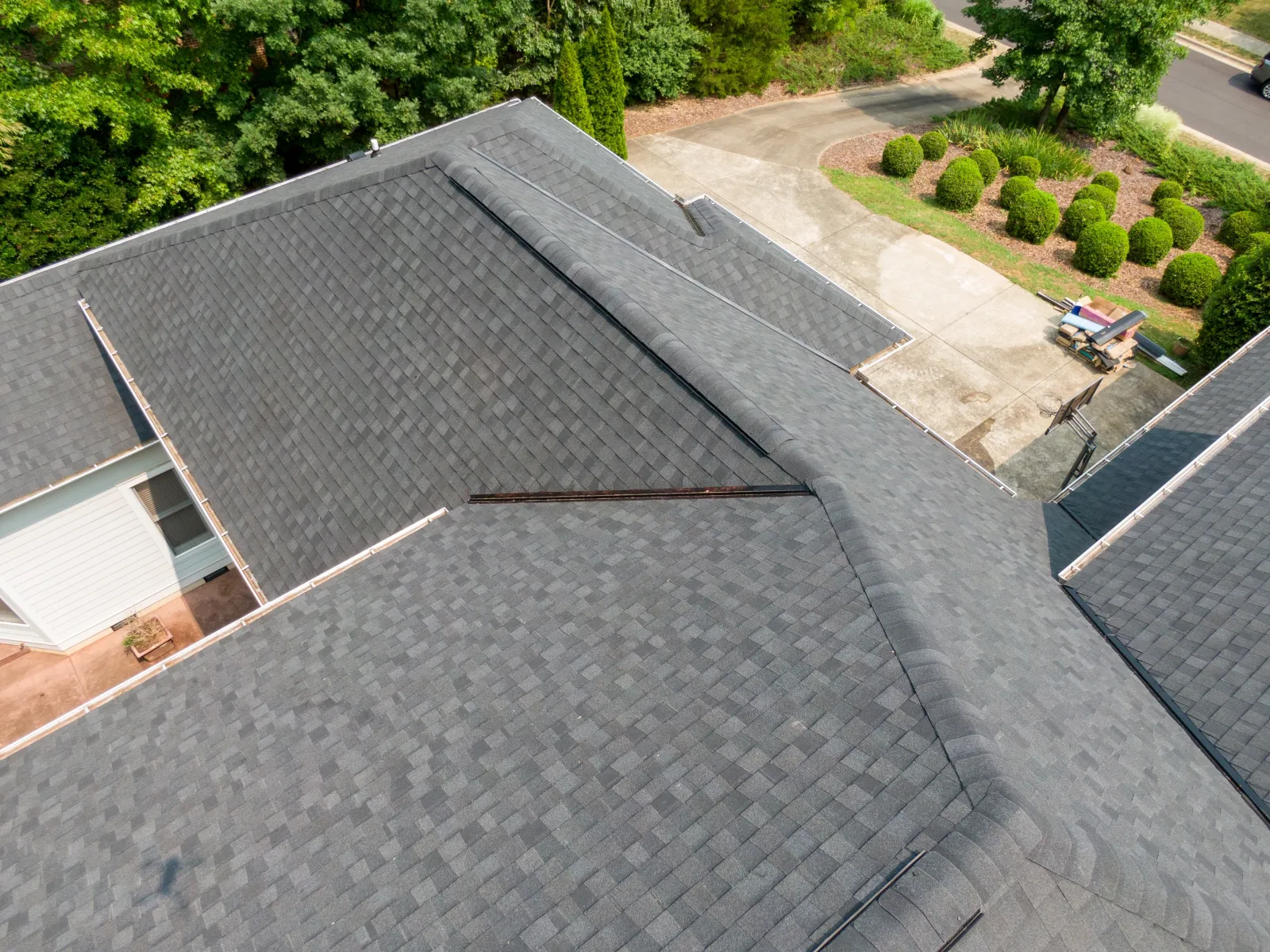 Aerial view of a large house roof with clean gray asphalt shingles surrounded by greenery and driveway.