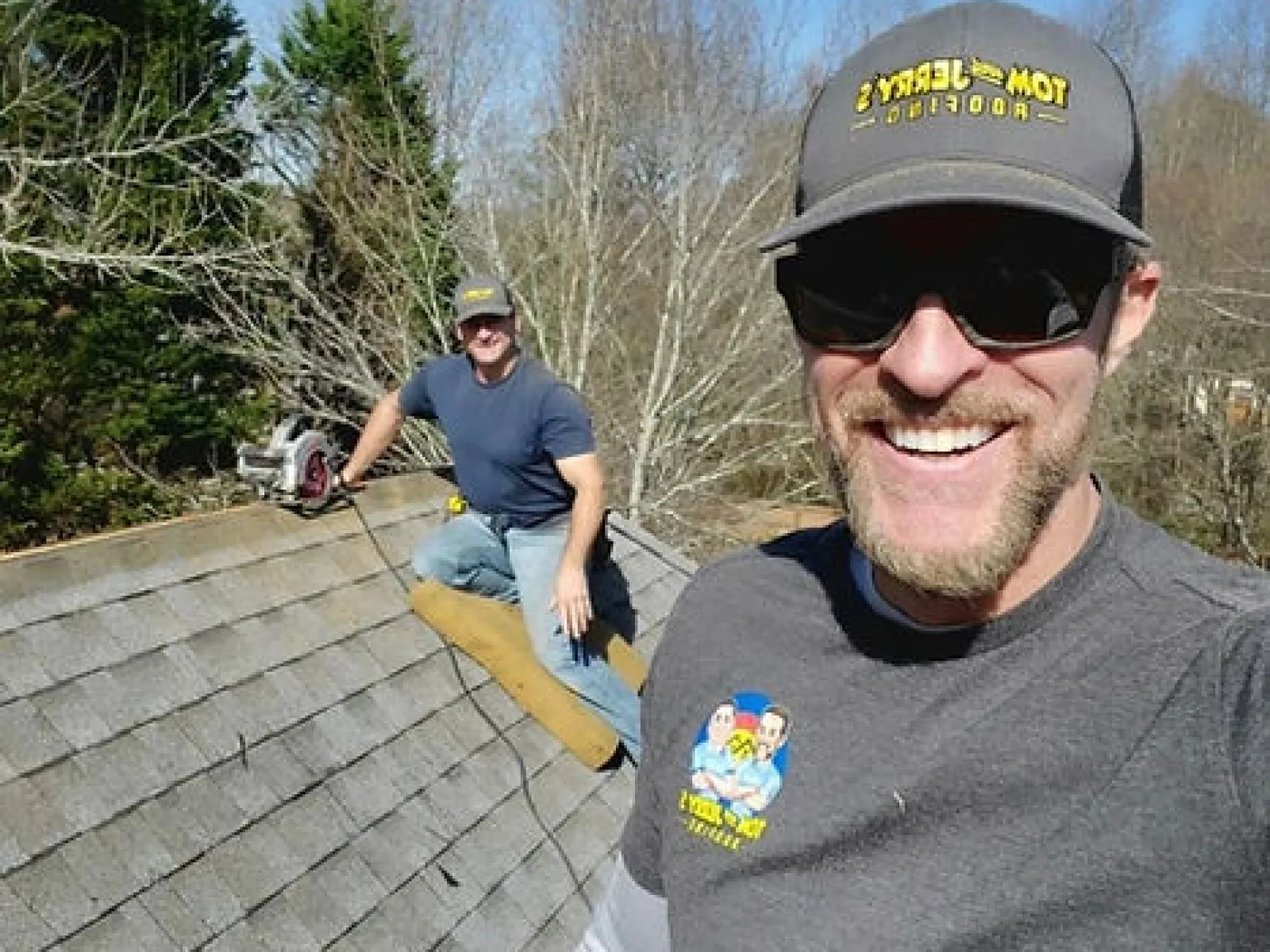Two men wearing Tom and Jerry's Pro Roofing hats working on a shingled roof on a clear day