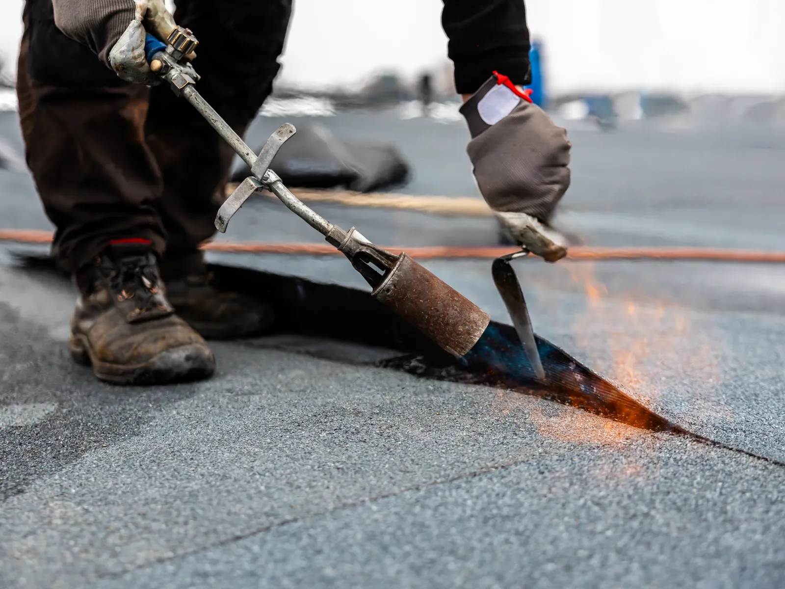 Worker using a torch to repair or install a flat roofing membrane with protective gloves and boots