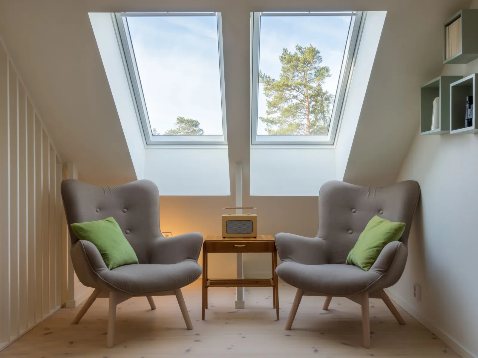 Cozy attic seating area with two gray armchairs, green pillows, wooden side table, and skylight windows.