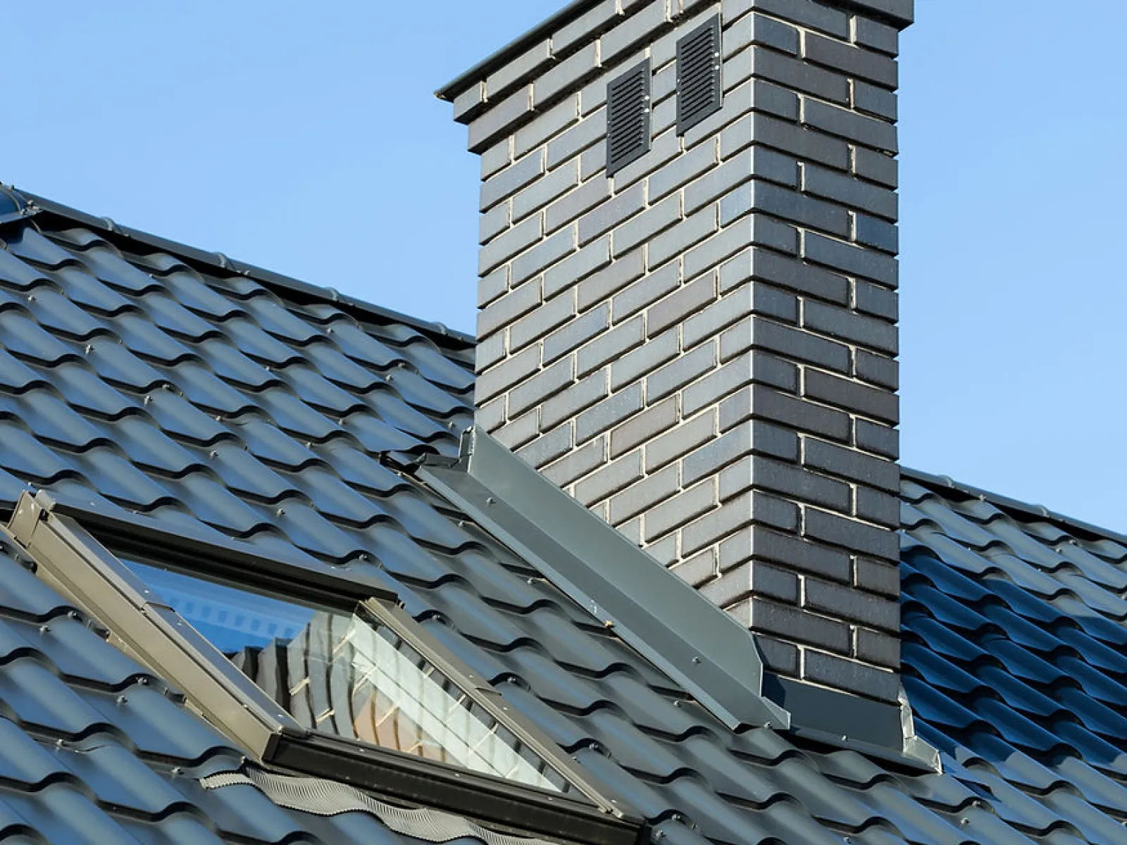 Close-up of dark metal roof with a skylight window and brick chimney against a clear blue sky.