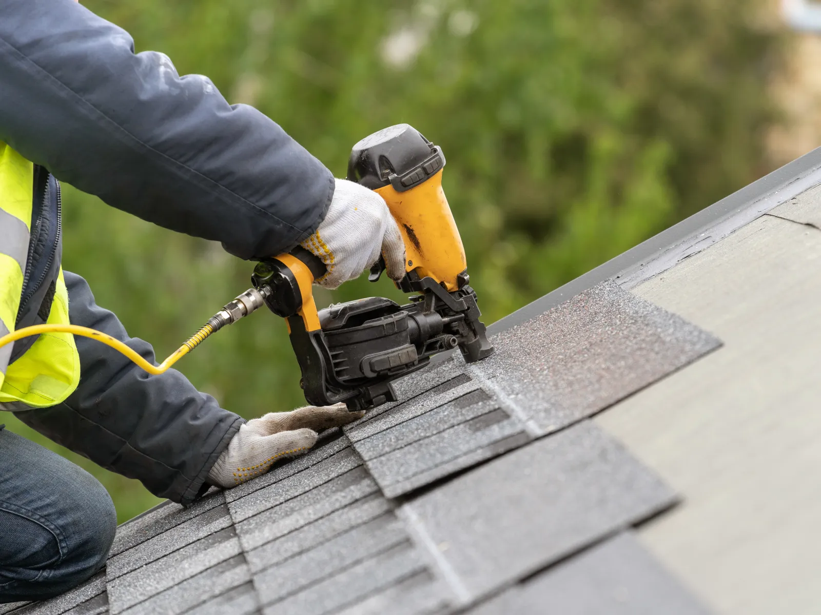 Construction worker wearing gloves and high-visibility jacket using nail gun to install roof shingles.
