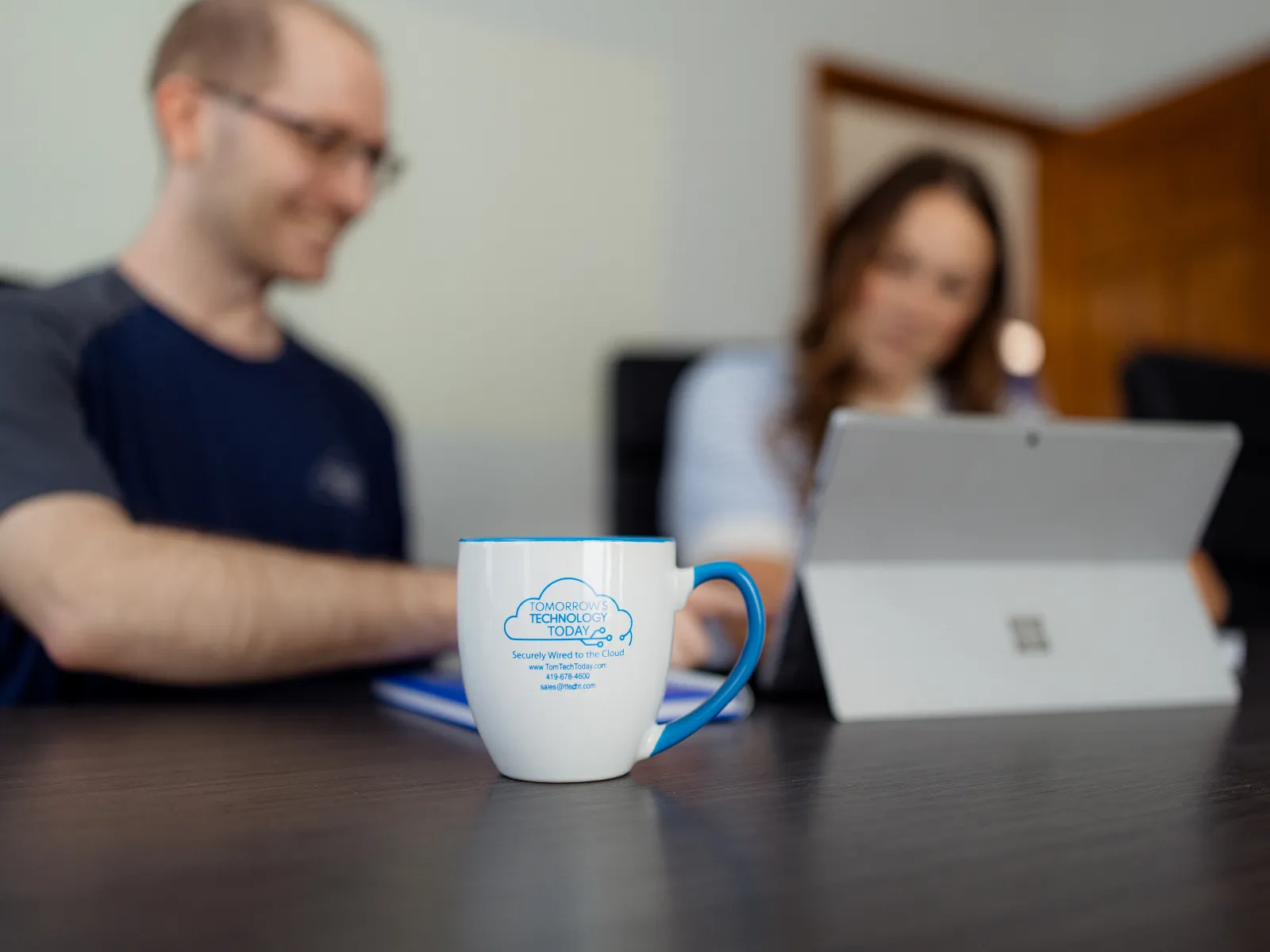 White coffee mug with blue handle and technology company logo on table, people working on a tablet in background.