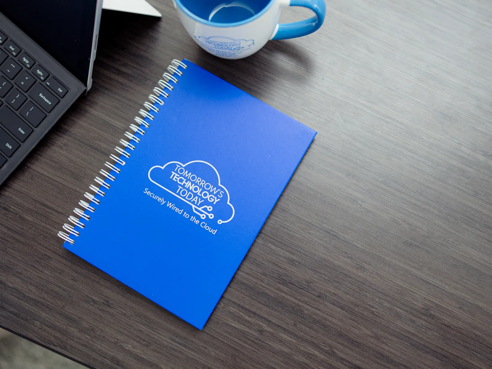 Blue spiral notebook with cloud technology logo on a wooden desk next to a coffee cup and laptop