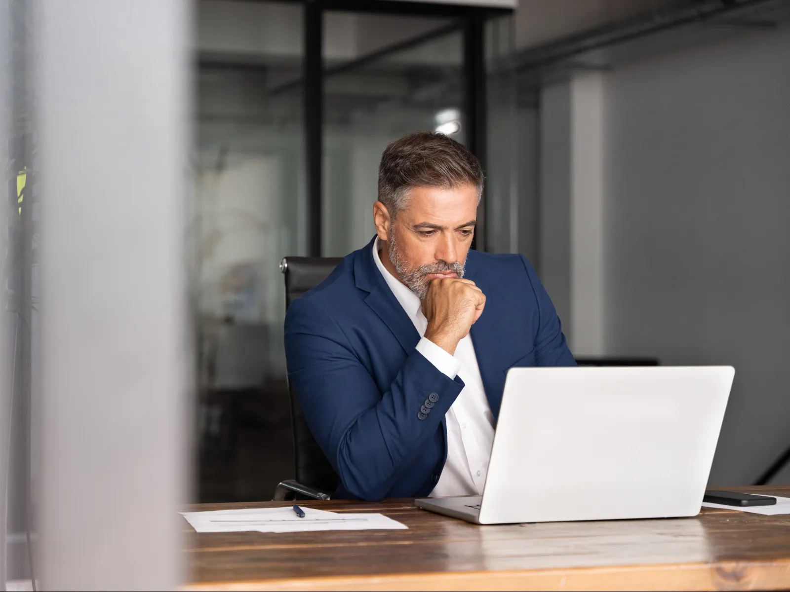 Businessman in a blue suit thoughtfully working on a laptop at a wooden desk in a modern office.