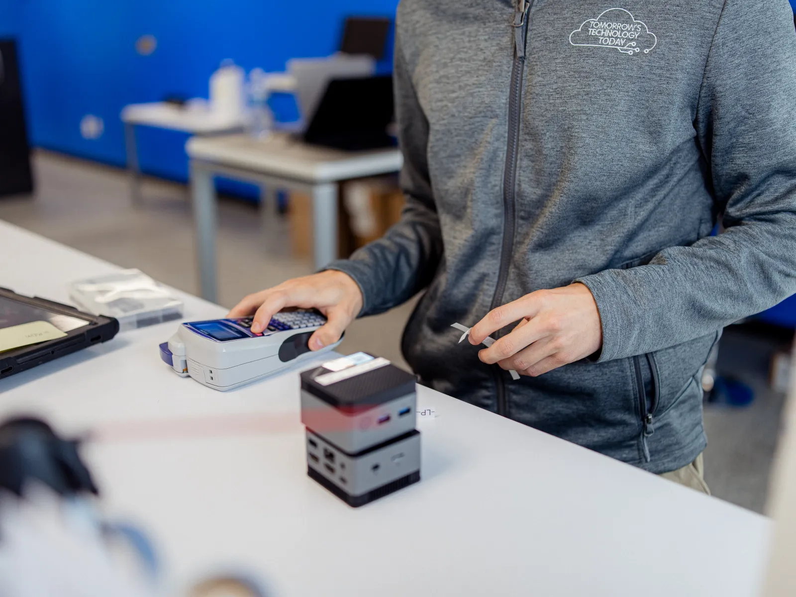 Person in gray jacket using handheld device and holding a small object at a white desk with tech equipment.