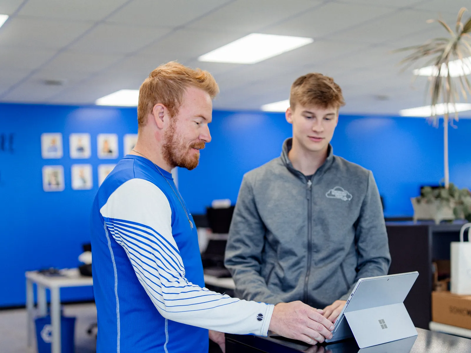 Two men collaborating in an office setting using a tablet on top of a black filing cabinet against a blue wall.