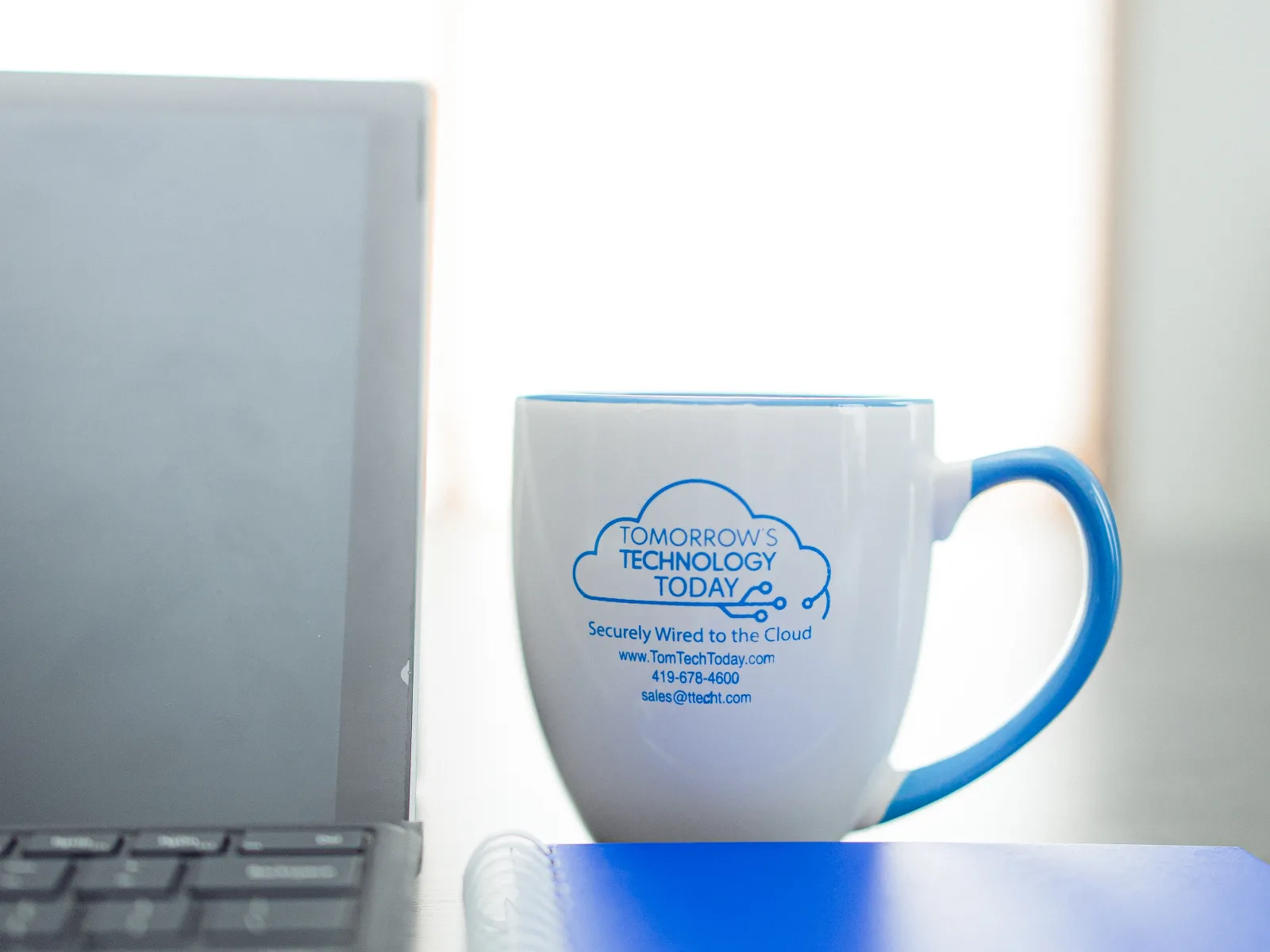 White coffee mug with blue handle and tech company logo next to laptop and blue notebook on desk.