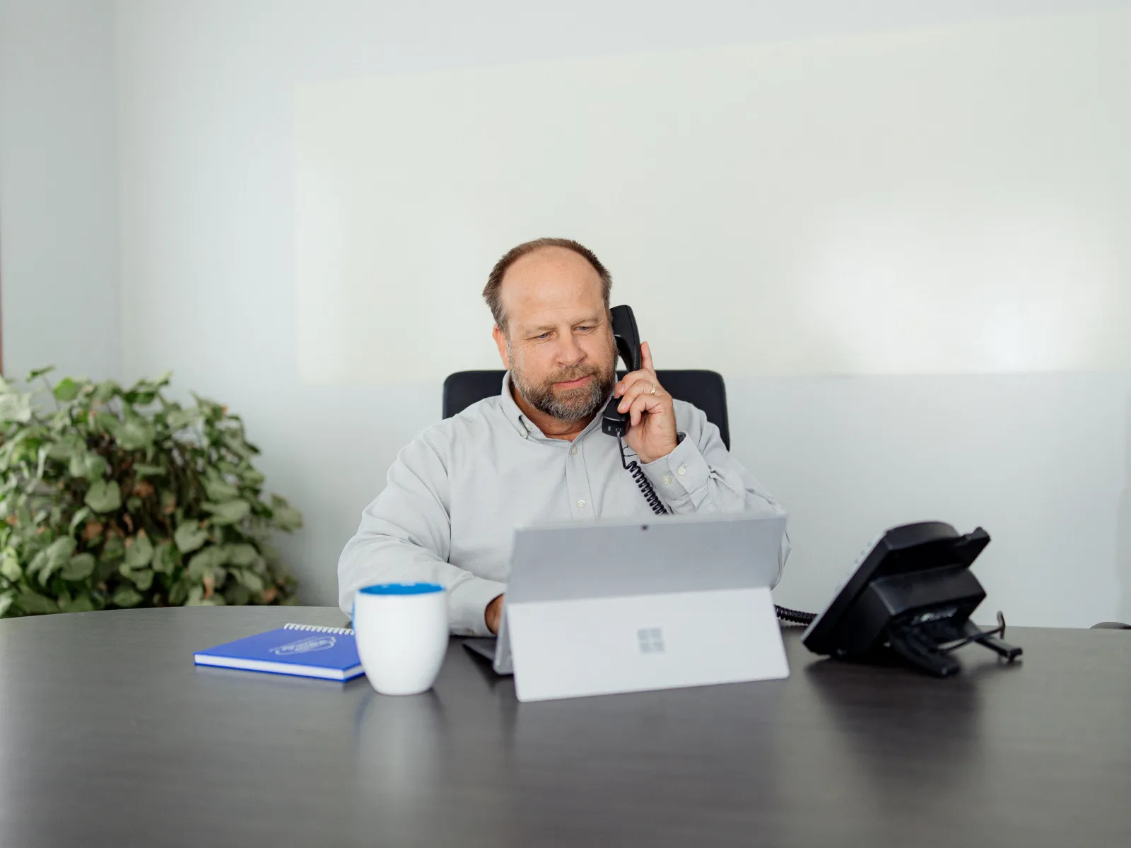 Man in office using a tablet while talking on a landline phone, with a mug and notebook on the desk