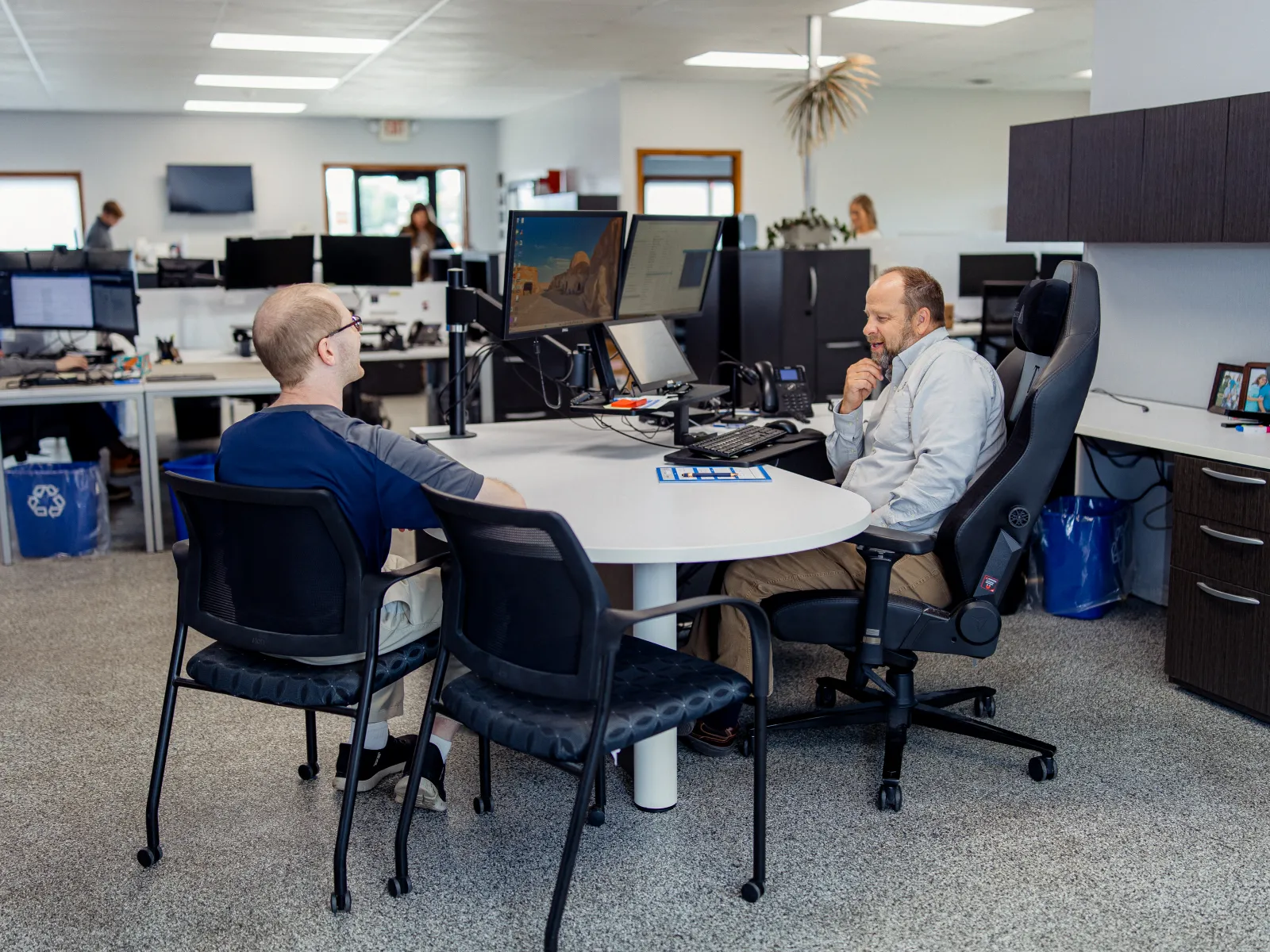 Two men having a discussion at a round table with monitors in a modern office workspace.