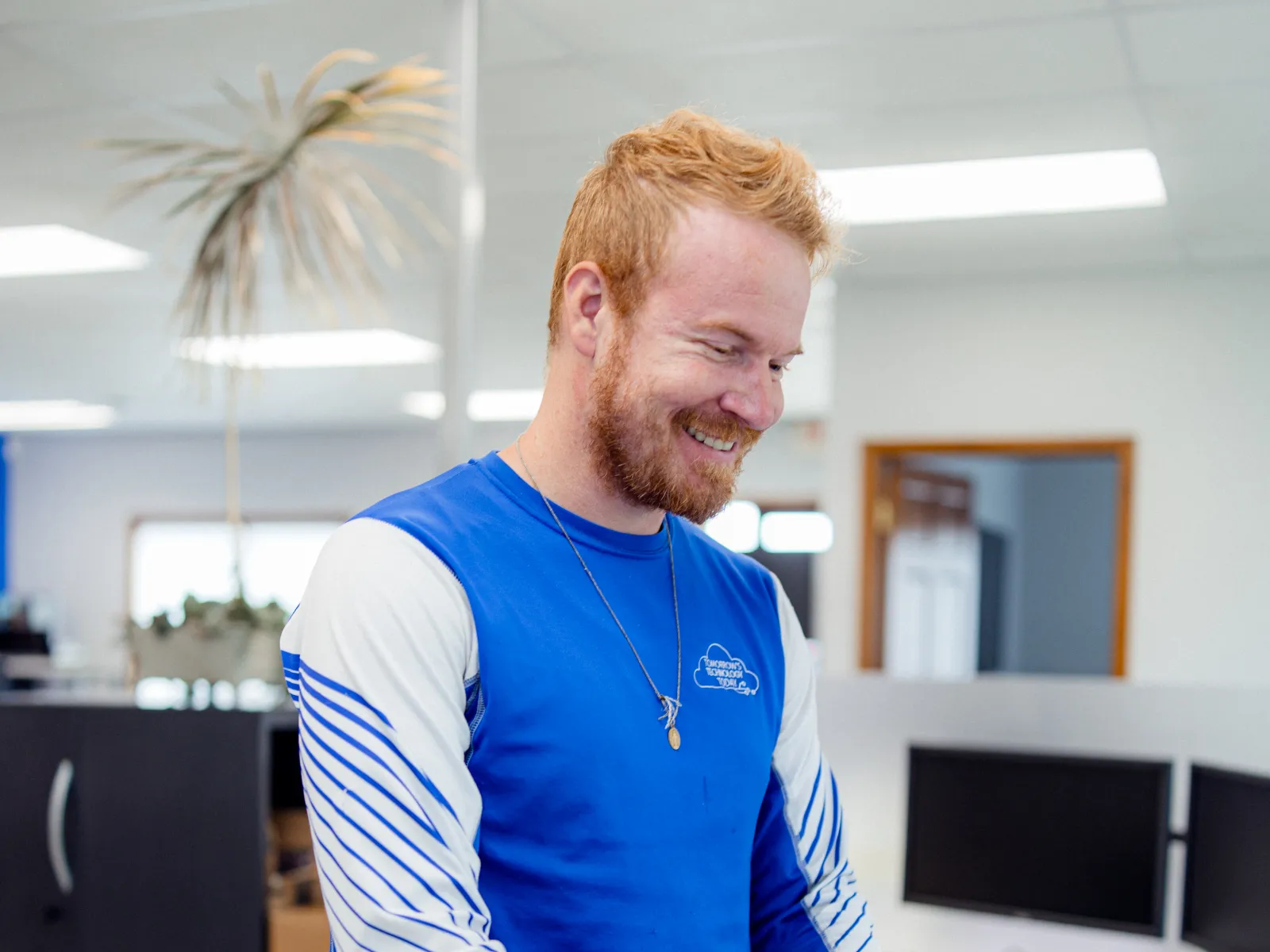 Smiling man in blue and white long-sleeve shirt typing on a laptop in a modern office space.