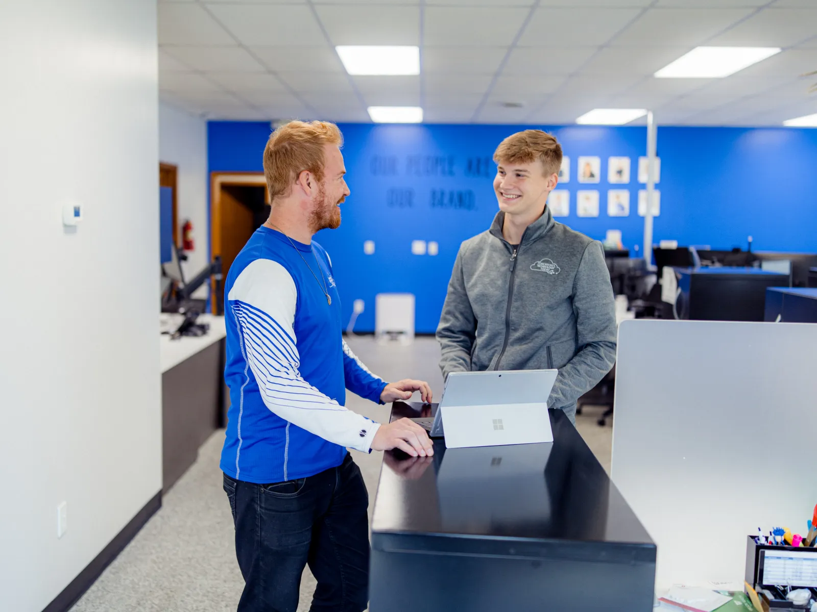 Two men smiling and talking at a modern office reception desk with a tablet and blue wall background.