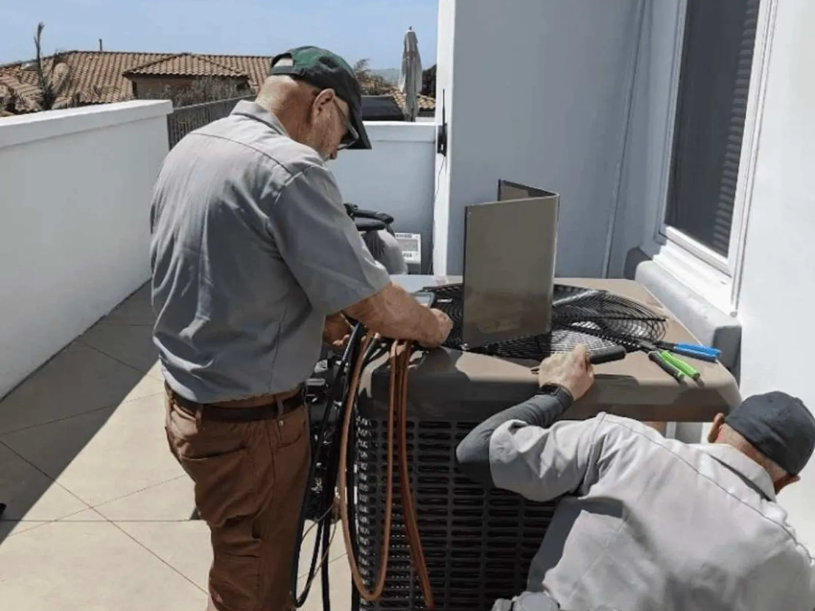 Two HVAC technicians repairing an air conditioning unit outside a residential building on a sunny day.