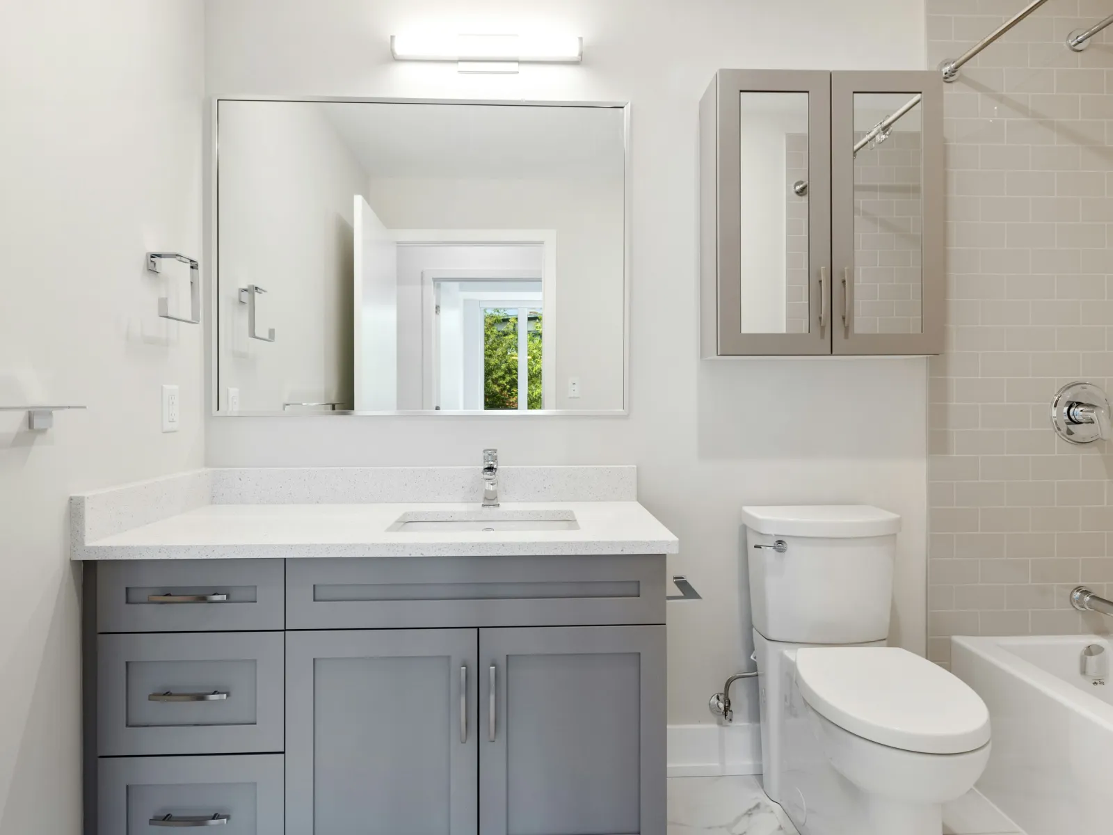 Modern bathroom with gray vanity, large mirror, white countertop, toilet, and tiled bathtub area under bright lighting