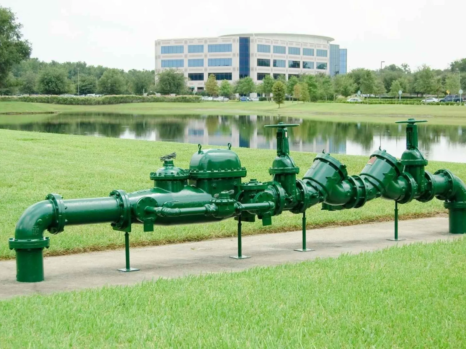Green industrial water pipe system with valves on grass near a pond and office building in the background