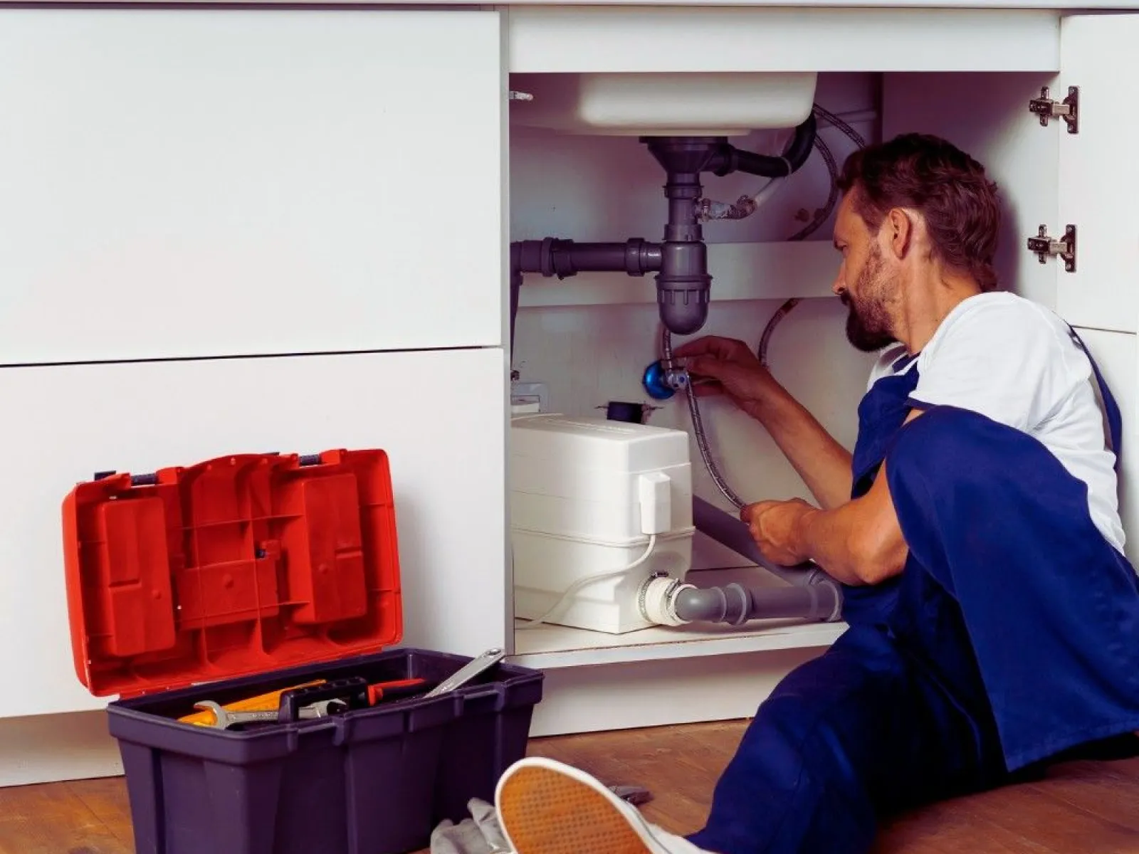 Plumber fixing pipes under kitchen sink with open toolbox on wooden floor nearby