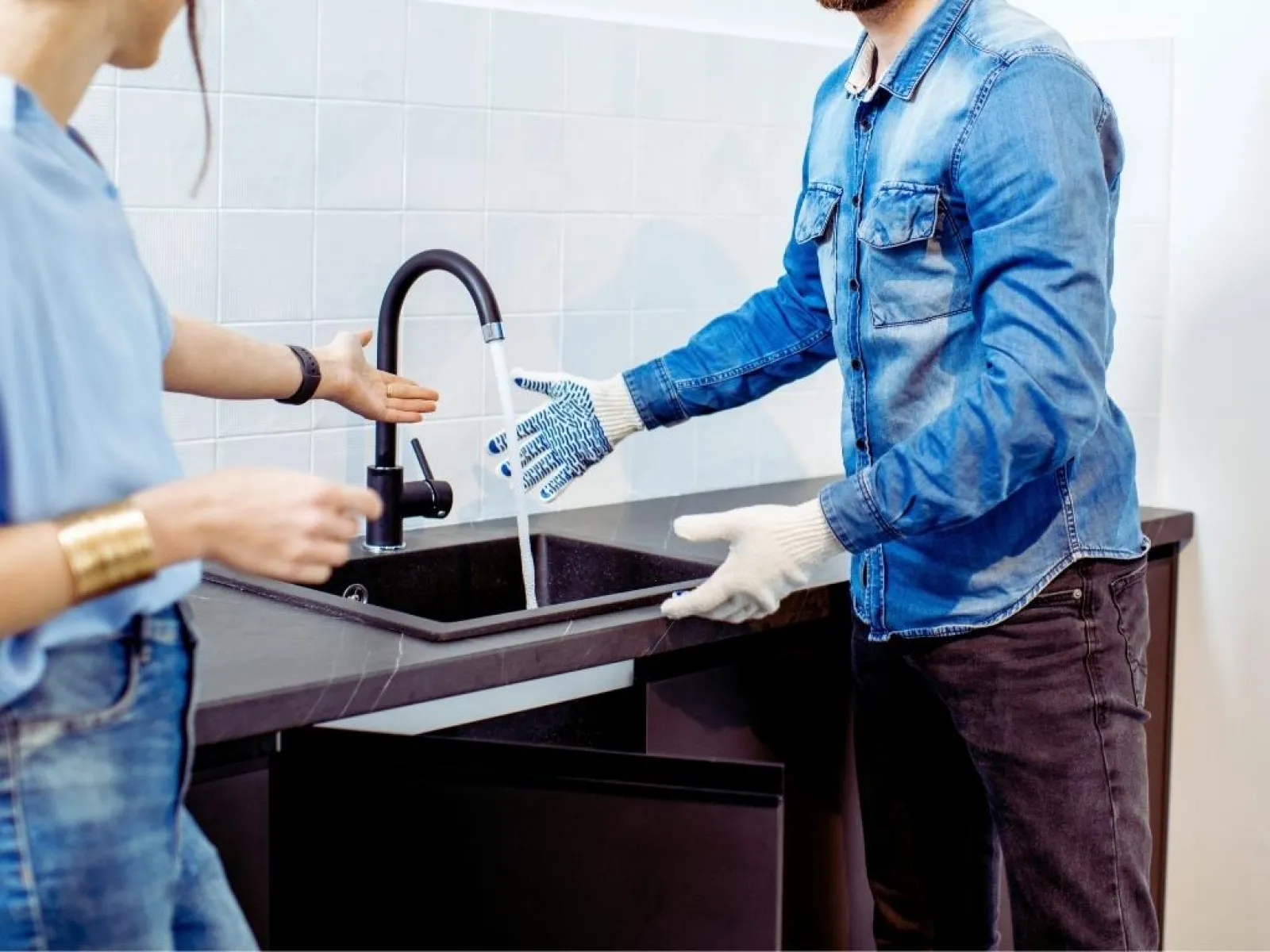 Two people testing a newly installed kitchen faucet with running water and open cabinet below.