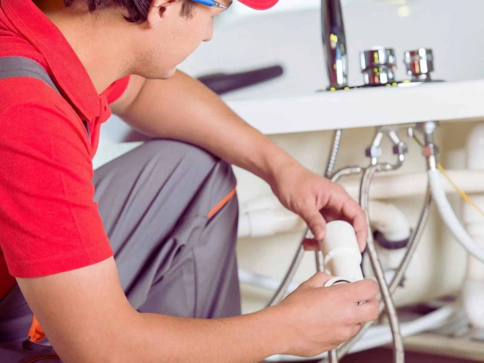 Plumber in red shirt and cap fixing pipe connections under a bathroom sink with a wrench and fittings.
