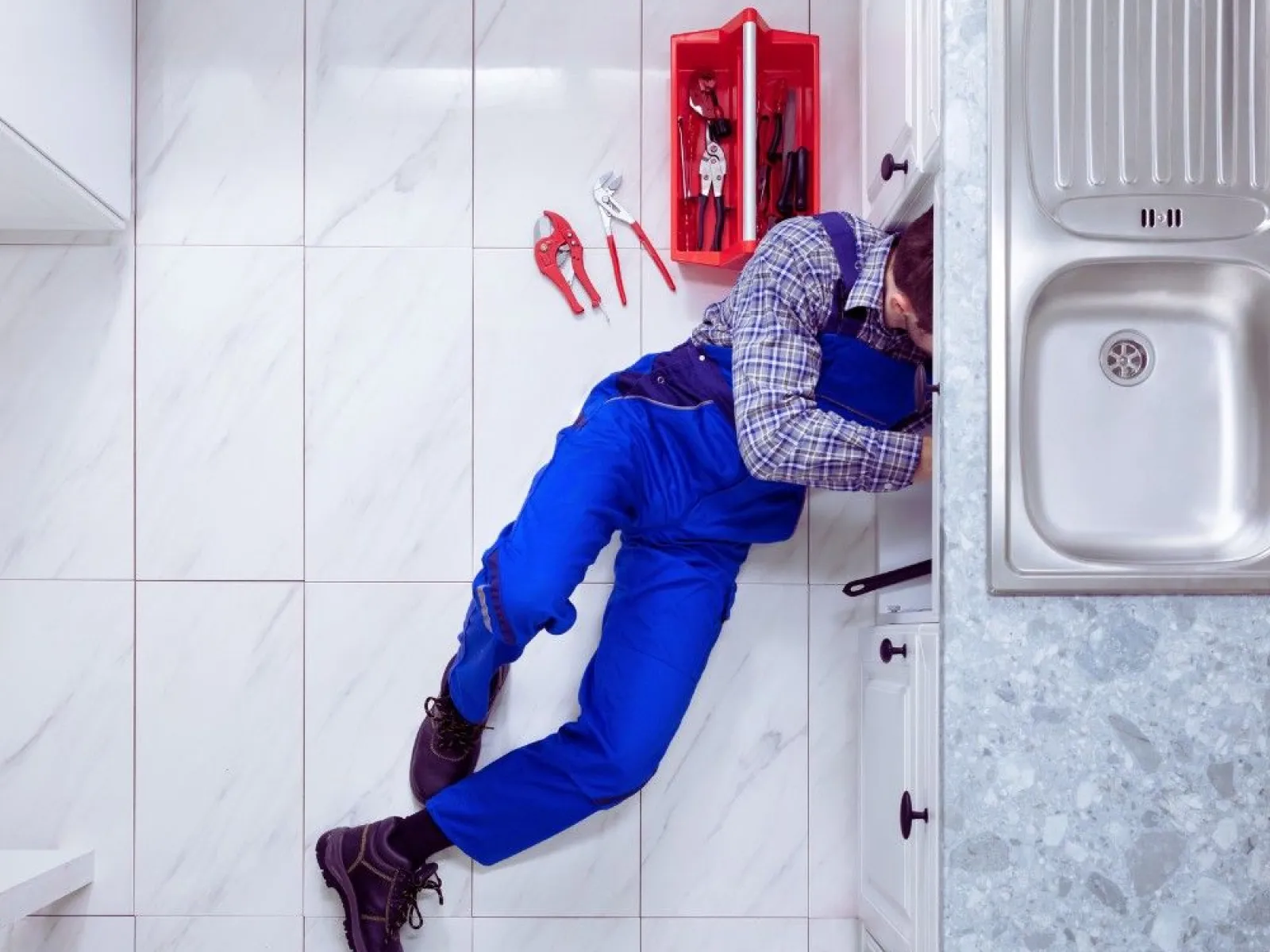 Plumber in blue overalls fixing kitchen sink under countertop with red toolbox and tools nearby on tiled floor